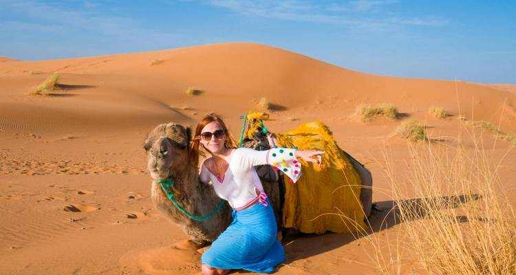2-Day Tour from Boumalne Dades showing a woman in a white blouse with colorful sleeves and blue skirt posing beside a saddled camel on golden sand dunes under a clear blue sky