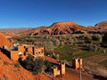 Landscape view of red clay buildings and green fields with rugged orange hills under a clear blue sky on a 2-Day Tour from Boumalne Dades.