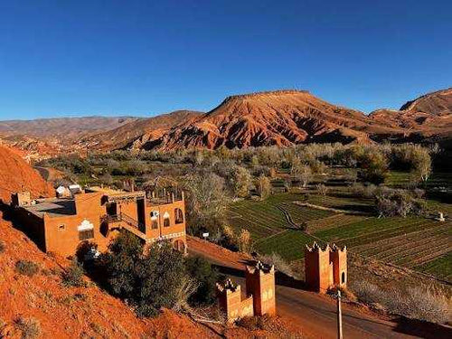 Landscape view of red clay buildings and green fields with rugged orange hills under a clear blue sky on a 2-Day Tour from Boumalne Dades.