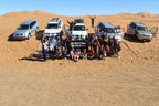 Group of tourists posing in front of five SUVs on orange sand dunes under a clear blue sky on a 2-Day Tour from Boumalne Dades