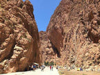 Narrow rocky Todra Gorge with towering orange cliffs under clear blue sky and tourists walking on dirt path