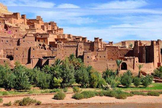 Ancient brown earthen kasbah with traditional Moroccan architecture surrounded by green palm trees under a blue sky in southeastern Morocco on 2-Day Tour from Marrakech