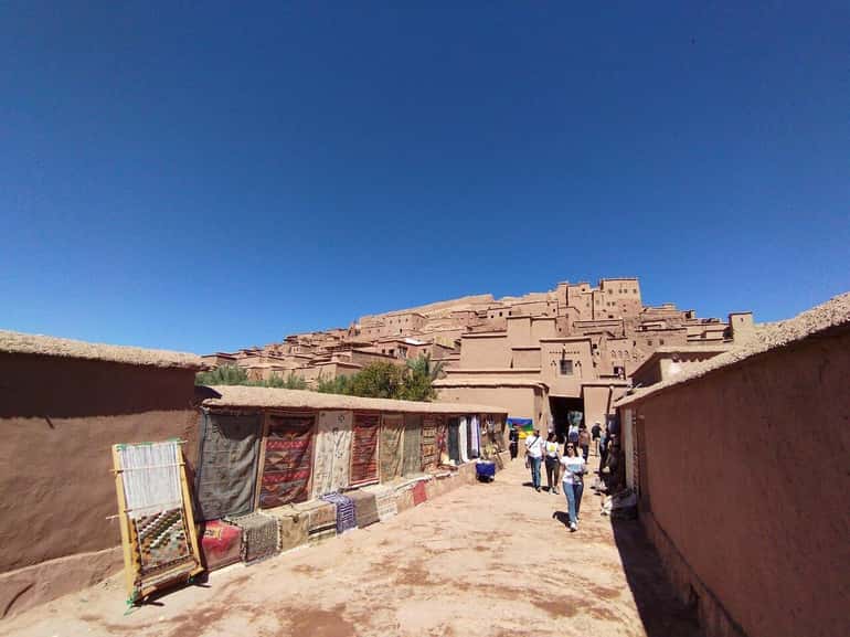 Ancient mud-brick kasbah village with narrow street lined by colorful woven rugs hanging on adobe walls under clear blue sky in desert