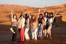 Group of seven women dressed in colorful summer clothes posing and smiling in the orange sand dunes of Merzouga during a 2-Day Tour from Marrakech