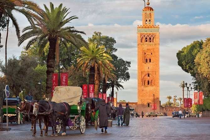Street view of traditional horse-drawn carriages and palm trees near a tall historic tower during a 2-Day Tour from Marrakech.