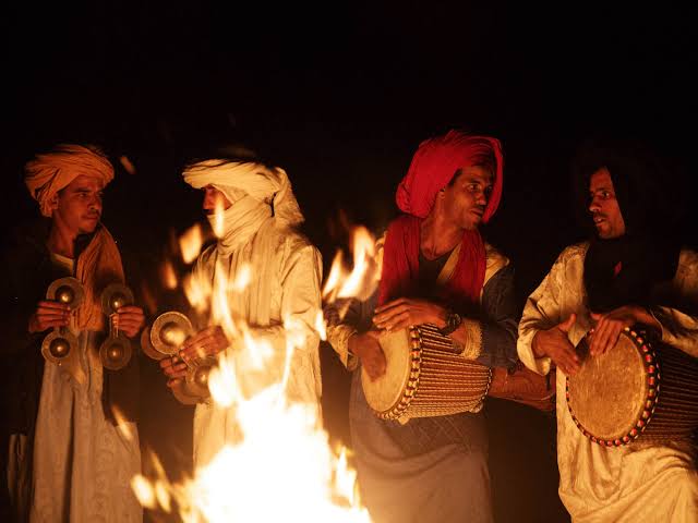 Traditional Moroccan musicians in colorful headscarves playing drums and cymbals around a desert campfire at night during a 2-Day Tour from Marrakech