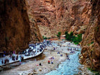 2-Day Tour from Marrakech showing rocky canyon with crowds of people, parked cars, and a flowing river along narrow pathway