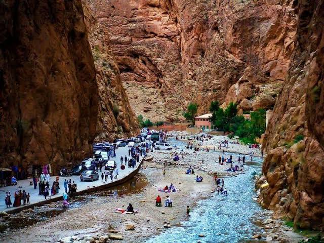 2-Day Tour from Marrakech showing rocky canyon with crowds of people, parked cars, and a flowing river along narrow pathway