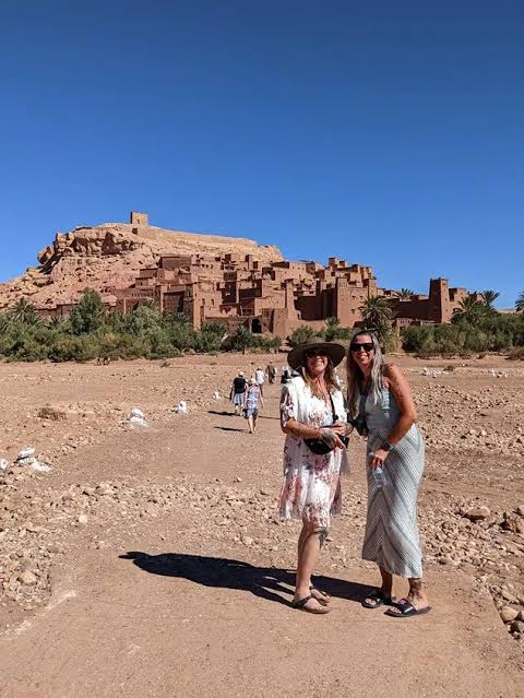 2-Day Tour from Marrakech featuring two women standing on a dirt path in front of a large, historic red clay kasbah under a clear blue sky
