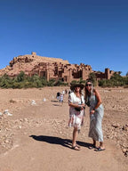 2-Day Tour from Marrakech featuring two women standing on a dirt path in front of a large, historic red clay kasbah under a clear blue sky