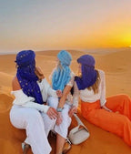 3 women in colorful scarves and flowing pants sitting on golden sand dunes at sunset during a 2-Day Tour from Marrakech in the Sahara Desert