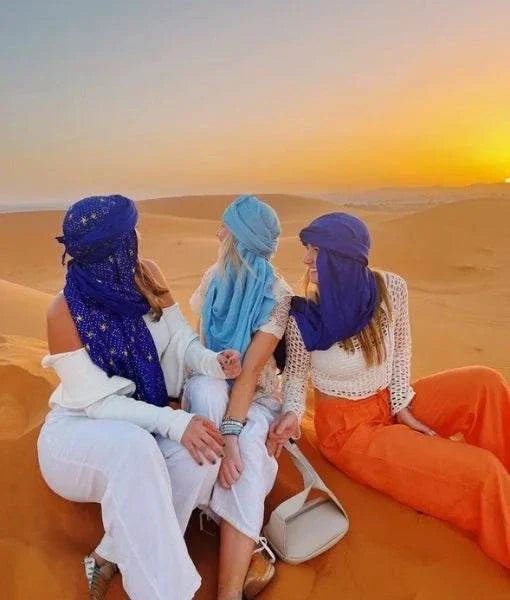 3 women in colorful scarves and flowing pants sitting on golden sand dunes at sunset during a 2-Day Tour from Marrakech in the Sahara Desert