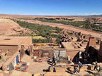 Mud-brick kasbah with colorful rugs and crafts displayed on walls and ground, overlooking a green valley in the 2 day tour from Merzouga to Marrakech.