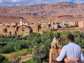 Landscape view of traditional brown clay buildings and lush green palm trees with rocky mountains in the background during a 2 day tour from Merzouga to Marrakech.