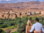 Landscape view of traditional brown clay buildings and lush green palm trees with rocky mountains in the background during a 2 day tour from Merzouga to Marrakech.