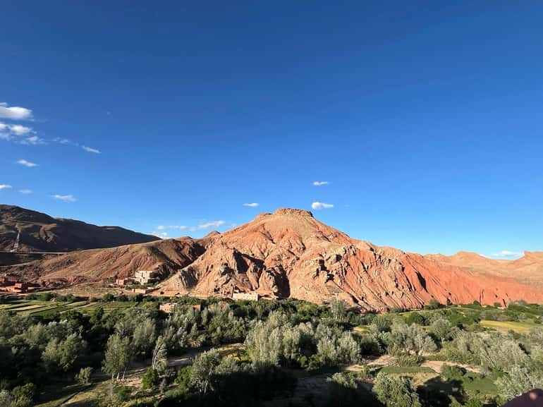 Landscape of rugged red and brown mountains with green forested valley under bright blue sky in 2 day tour from Merzouga to Marrakech.