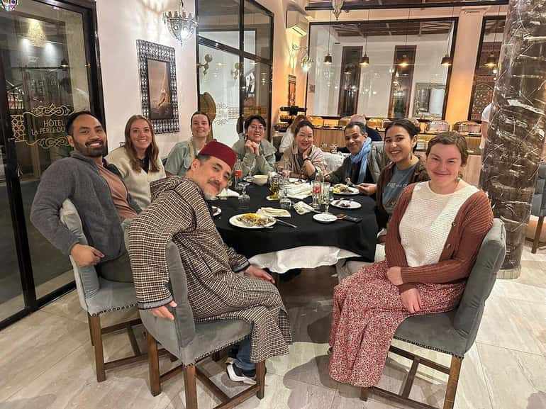 Group of people dining together around a black tablecloth-covered table with plates and drinks in a hotel restaurant, illustrating the 2 day tour from Merzouga to Marrakech experience.