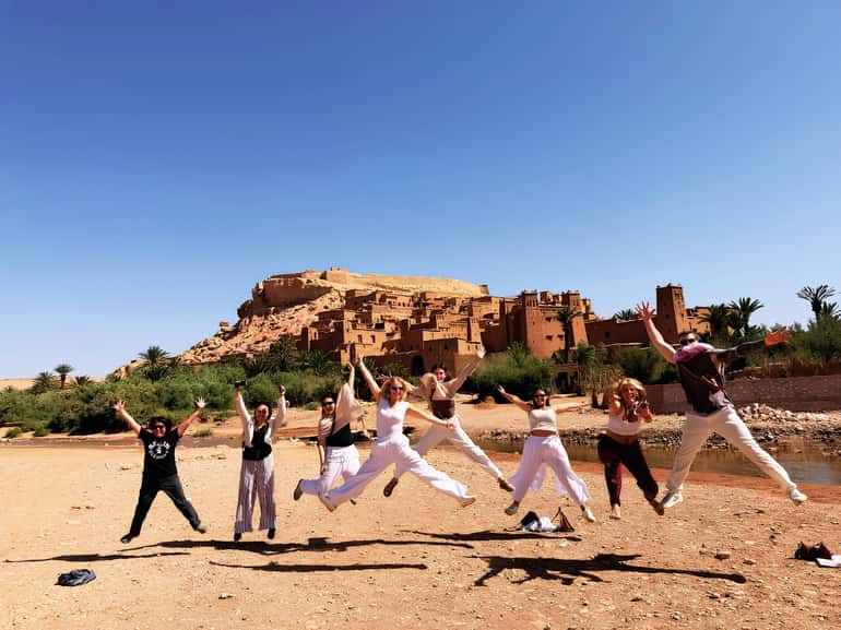 Group of tourists jumping joyfully on sandy ground with traditional brown clay buildings and lush greenery in the background on a 2 day tour from Merzouga to Marrakech