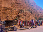 Outdoor market stalls with colorful textiles, scarves, and jewelry displayed against large orange rock cliffs, showcasing a vibrant scene from a 2 day tour from Merzouga to Marrakech.