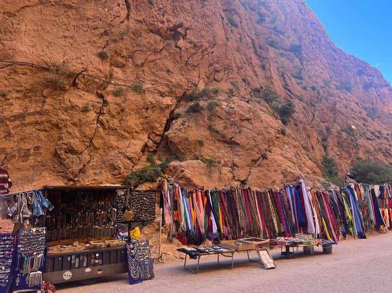 Outdoor market stalls with colorful textiles, scarves, and jewelry displayed against large orange rock cliffs, showcasing a vibrant scene from a 2 day tour from Merzouga to Marrakech.