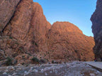 Rock canyon landscape with towering reddish-brown limestone cliffs and a rocky riverbed under a clear blue sky, showcasing the 2 day tour from Merzouga to Marrakech.