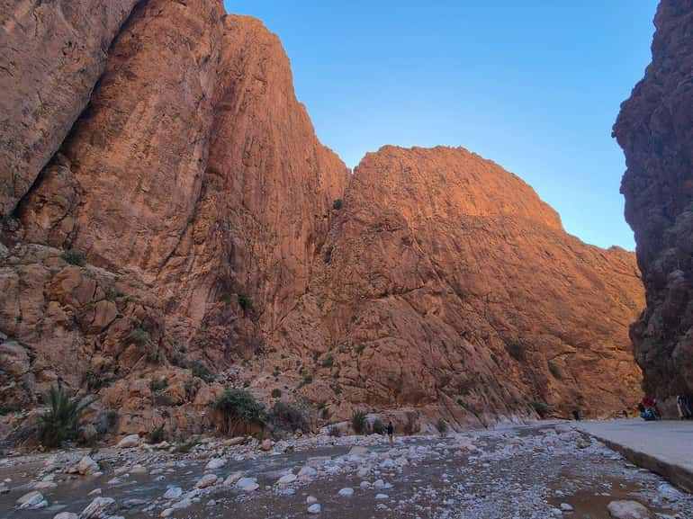 Rock canyon landscape with towering reddish-brown limestone cliffs and a rocky riverbed under a clear blue sky, showcasing the 2 day tour from Merzouga to Marrakech.