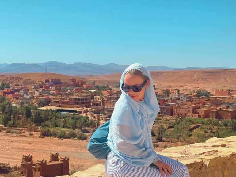 Woman wearing light blue shawl, sunglasses, and striped pants sitting on stone with desert town and hills in background, 2 day tour from Merzouga to Marrakech.