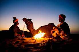 Desert camel and two men wearing traditional Moroccan clothing sitting by a campfire at sunset on the sand dunes during a 3 Days Tour from Fes to Merzouga.