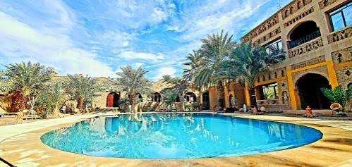 Outdoor swimming pool with clear blue water surrounded by palm trees and a multi-story rustic beige building under a partly cloudy sky during 3 Days Tour from Fes to Merzouga.