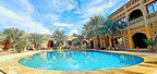 Outdoor swimming pool with clear blue water surrounded by palm trees and a multi-story rustic beige building under a partly cloudy sky during 3 Days Tour from Fes to Merzouga.