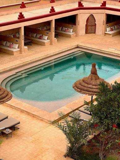 Outdoor Moroccan-style swimming pool with beige stone tiles, thatched umbrella, and cushioned seating alcoves, perfect for relaxing during a 3 Days Tour from Fes to Merzouga.