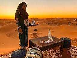 Desert tour setup with a person in traditional Moroccan attire holding a teapot, serving tea on a low wooden table with cups, cushions, and a rug on sand dunes at sunset, highlighting the 3 Days Tour from Fes to Merzouga.