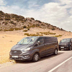 Gray Ford passenger van and black SUV parked on a roadside in a rocky desert landscape during the 3 Days Tour from Fes to Merzouga.