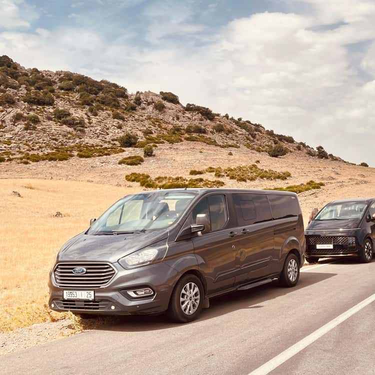 Gray Ford passenger van and black SUV parked on a roadside in a rocky desert landscape during the 3 Days Tour from Fes to Merzouga.