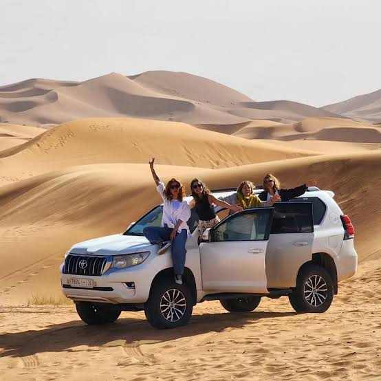 White Toyota SUV with four women posing joyfully in front of large desert sand dunes during a 3 Days Tour adventure