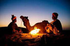 3 Days Tour featuring two men in traditional Moroccan attire sitting by a campfire with a camel in desert sand at sunset.