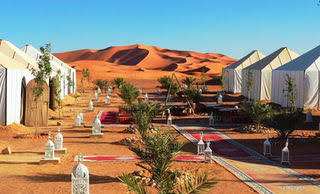 Luxury desert camp with white canvas tents, colorful rugs, palm plants, and lanterns set against Erg Chebbi dunes during 3 Days Tour