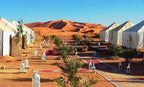 Luxury desert camp with white canvas tents, colorful rugs, palm plants, and lanterns set against Erg Chebbi dunes during 3 Days Tour
