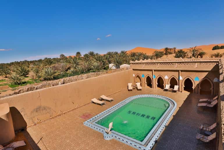 Desert riad with traditional Moroccan architecture and an outdoor swimming pool surrounded by palm trees and sand dunes on a 4-day desert tour