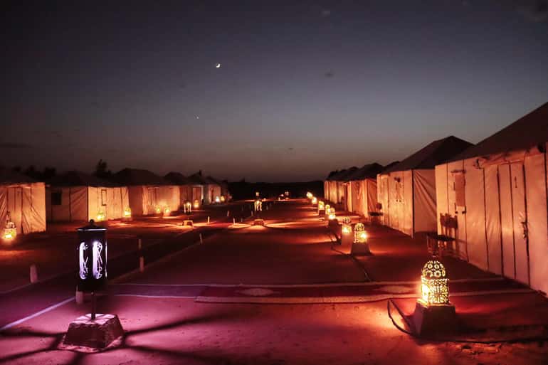 Desert camp at night with illuminated tents and decorative lanterns lining a sandy pathway under a crescent moon sky, perfect for a 4-day desert tour
