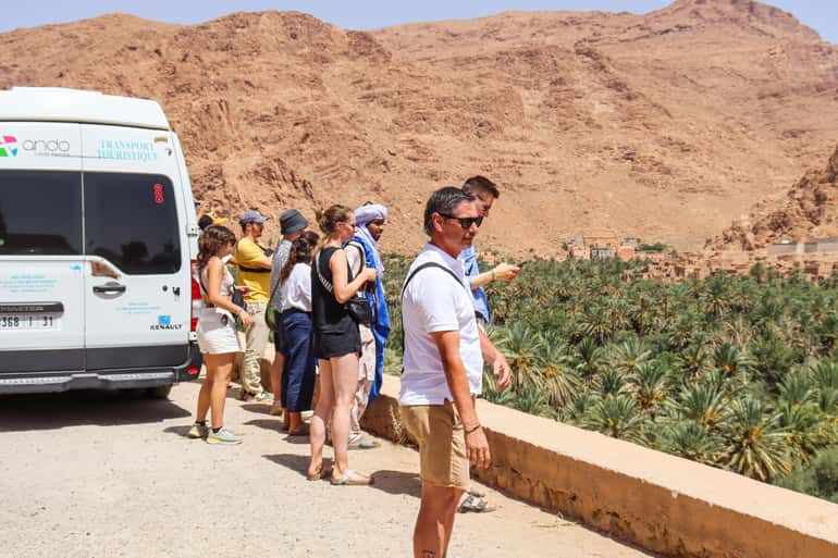 Group of tourists in casual summer clothing standing near white tourist van, overlooking green palm oasis against rocky desert hills on 4-day desert tour.