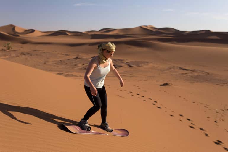 Person wearing white tank top and black pants sandboarding on orange desert sand dunes during 4-day desert tour