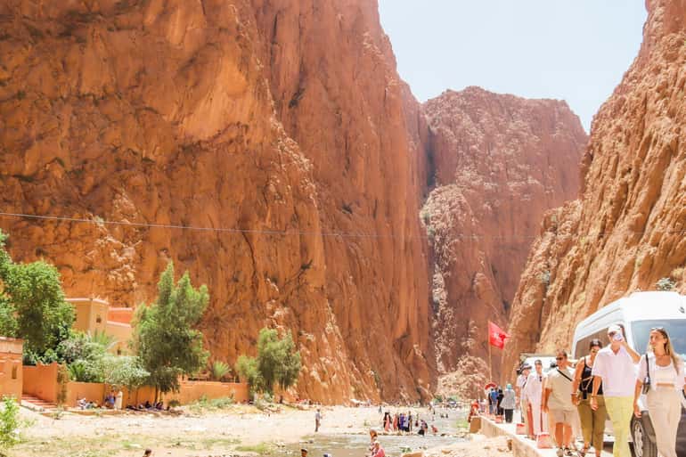 4-day desert tour showing tourists walking between towering orange-red rocky cliffs with scattered greenery and vehicles nearby