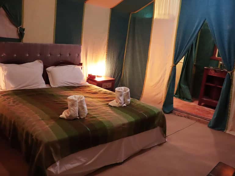 Bed with green and brown striped blanket and white pillows in a tented desert camp room softly lit with bedside lamps for 4-day desert tour