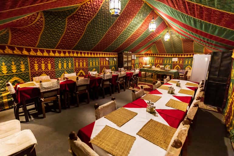 4-day desert tour dining tent with long tables covered in red and white cloths, wicker placemats, and traditional colorful fabric decor