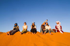 Group of six diverse travelers sitting on bright orange desert dunes under a clear blue sky during a 4-day desert tour
