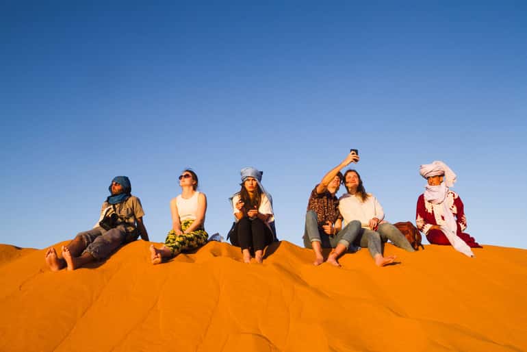 Group of six diverse travelers sitting on bright orange desert dunes under a clear blue sky during a 4-day desert tour