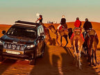 Black Toyota 4x4 vehicle parked on orange desert sand dunes alongside group of camels with riders in colorful clothing