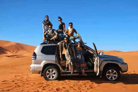 Silver 4x4 SUV parked on orange desert sand with five people posing around and on top under clear blue sky 4x4 Rental in Merzouga