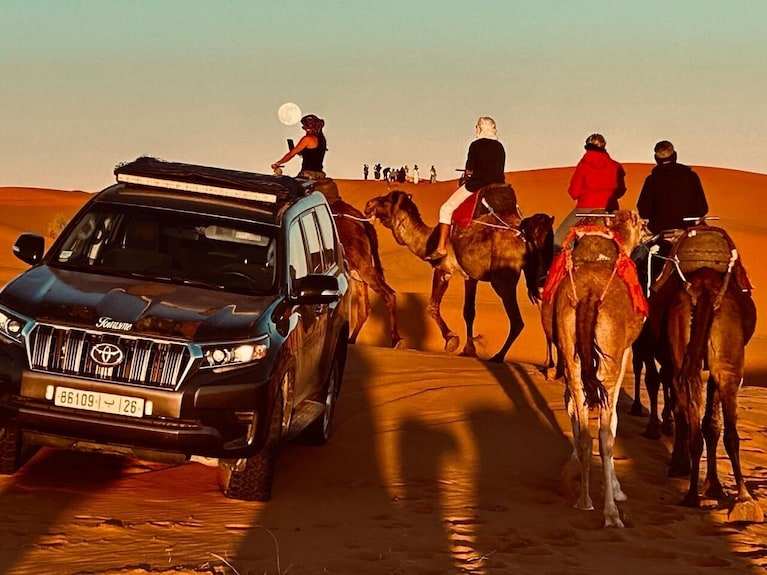 4x4 Transfer black Toyota SUV driving on golden sand dunes alongside riders on camels in desert landscape